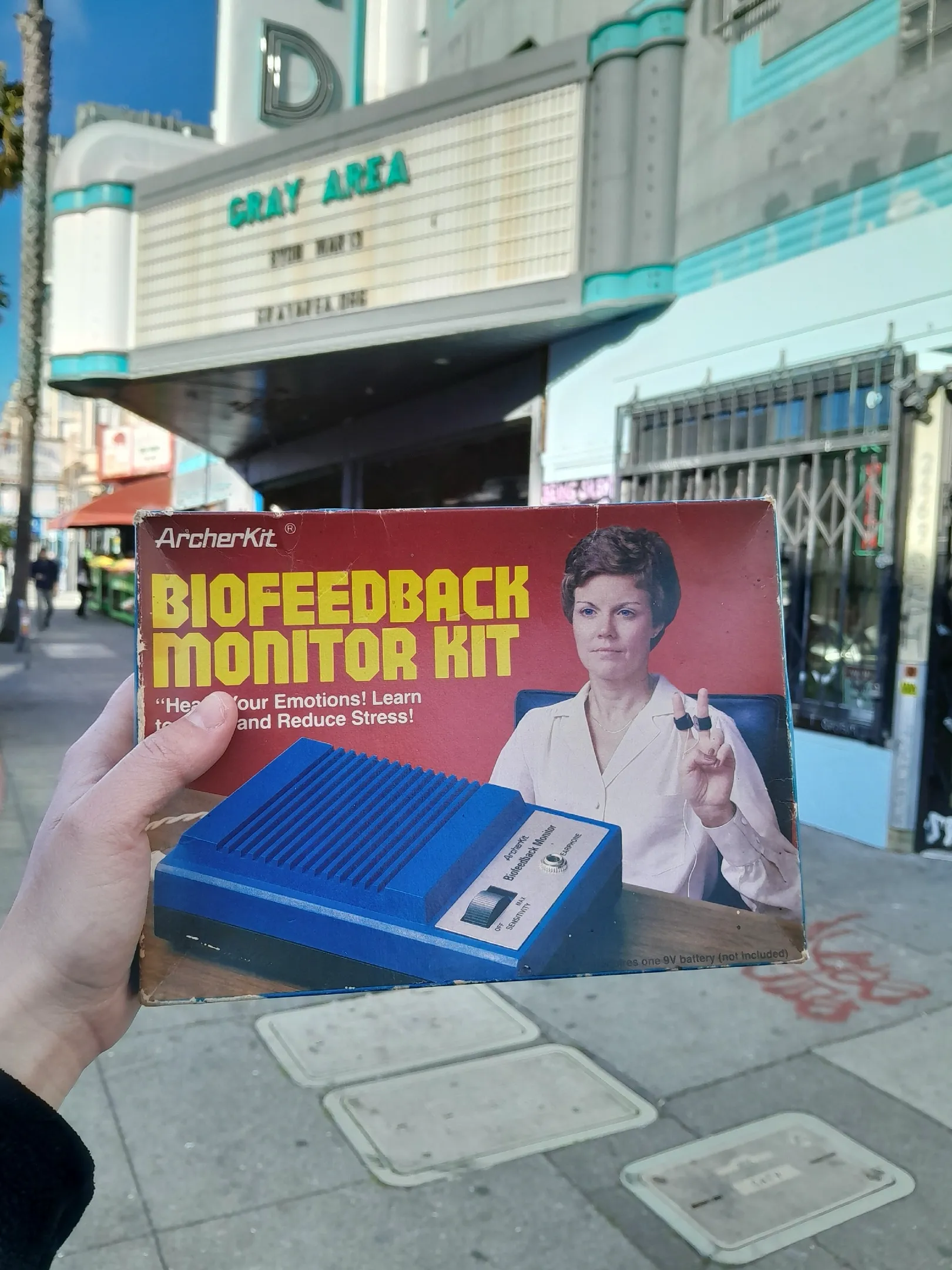 A person holds a box of a biofeedback monitor, showcasing its packaging and features in front of the Gray Area building.