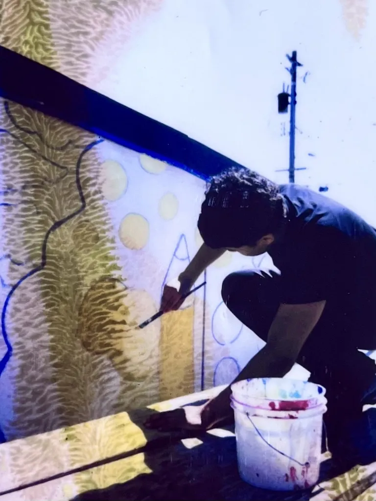 A person crouches while painting a mural on an outdoor wall, holding a brush and a paint bucket, with abstract shapes visible on the wall.