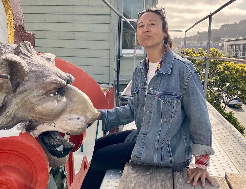 Julia in a denim jacket sits next to a large, decorative lion sculpture on a rooftop with buildings and trees in the background.